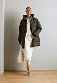 Donkergroene pufferjack met een rits- en knoopsluiting, gecombineerd met een witte blouse en een lange witte rok, terwijl ze een tote bag vasthoudt.