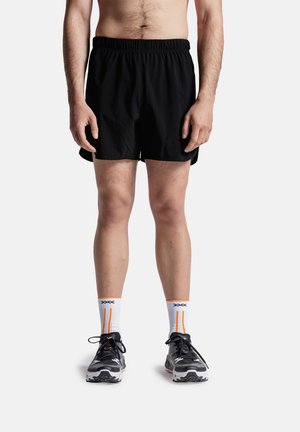 Man draagt zwarte sportshorts, witte sportsokken met oranje strepen en zwarte hardloopschoenen, staand tegen een witte achtergrond.