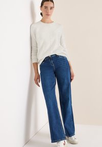Hellgrauer, langärmliger Pullover mit gerippten Bündchen, kombiniert mit blauen weit geschnittenen Jeans. Das Model steht vor einem hellen Hintergrund.