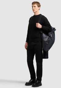 Jeune homme portant un pull noir, un pantalon et des chaussures, portant une doudoune bleu marine sur l'épaule sur un fond clair uni.
