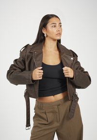 Giacca cropped in finta pelle marrone con chiusura a zip, spalline e dettagli metallici. Indossata sopra una canotta nera e pantaloni beige.