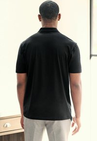 Polo negro de manga corta con una textura suave, que cuenta con un cuello clásico y un dobladillo recto. Llevado con pantalones gris claro.
