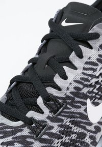 Nike sportssko med et sort og gråt mønstret mesh-overdel, teksturerede snørebånd og en hvid logo på siden. Har forstærkede snørehuller.