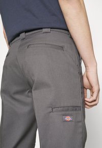 Gros plan sur un pantalon de travail gris avec des poches arrière et latérales, un patch de la marque Dickies sur la cuisse, porté avec une chemise bleu marine.