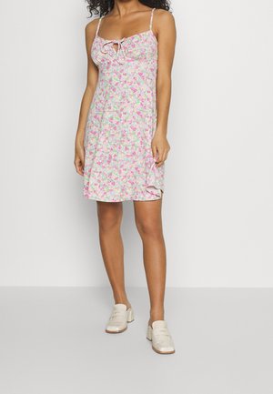 Femme portant une robe courte sans manches à fleurs avec de fines bretelles et des mocassins blancs à petit talon, debout devant un fond clair uni.