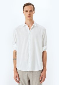 Camisa blanca de lino con botones y mangas enrolladas, que presenta un corte relajado y una tela texturizada. Material suave y ligero con un ligero brillo.