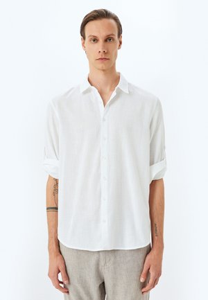 Camisa blanca de lino con botones y mangas enrolladas, que presenta un corte relajado y una tela texturizada. Material suave y ligero con un ligero brillo.
