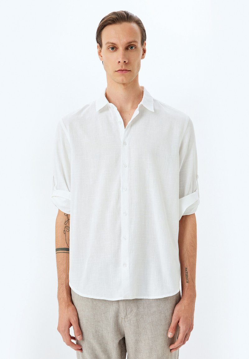 Camisa blanca de lino con botones y mangas enrolladas, que presenta un corte relajado y una tela texturizada. Material suave y ligero con un ligero brillo.