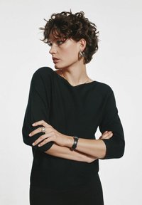 Czarny, długi sweter z dzianiny z szerokim dekoltem i luźnym krojem. Modelka nosi akcesoria, w tym pierścionek i czarny zegarek.