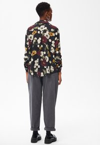 Personne avec les cheveux courts portant des boucles d'oreilles en créole, une blouse florale noire à pois blancs, un pantalon gris ample, des chaussettes noires et des mocassins noirs, debout.