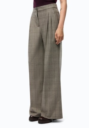 Donna con pantaloni a gamba larga a quadri con pieghe frontali e scarpe platform marrone scuro, in piedi davanti a uno sfondo semplice.