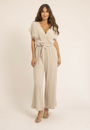 Jumpsuit beige enroulé en tissu texturé. Caractéristiques : manches courtes, taille ceinturée et pantalon large. Porté avec des talons hauts à lanières.
