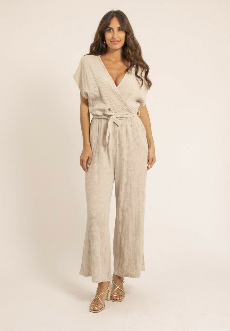 Jumpsuit beige enroulé en tissu texturé. Caractéristiques : manches courtes, taille ceinturée et pantalon large. Porté avec des talons hauts à lanières.