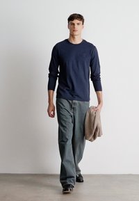 Mornarska majica s dolgimi rokavi z majhnim logotipom, v kombinaciji s širokimi sivimi kavbojkami. Drži svetlo rjavo pulover in nosi črne čevlje, na nevtralni ozadju.