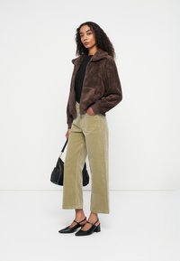 Bruine faux fur jas met ritssluiting, olijfgroene corduroy broek met wijde pijpen en zwarte slingback hakken. Model draagt een zwarte schoudertas.