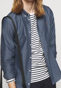 Donkerblauw overhemd van gladde stof, gedragen over een wit T-shirt met marineblauwe strepen; voorzien van een kleine geborduurde accenten.