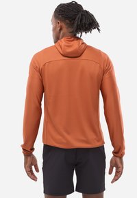 Haut à capuche de sport orange à manches longues avec un motif texturé, capuche réglable et poignets ajustés, associé à un short noir.
