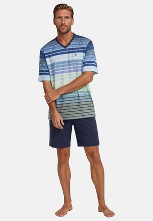 Mann steht barfuß da, trägt ein blau-grün gestreiftes V-Ausschnitt-Shirt und dunkelblaue Shorts, die Arme vor dem Oberkörper verschränkt.