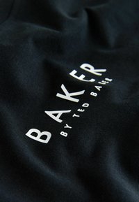 Tissu noir présentant un texte blanc en relief : « BAKER » en lettres grasses et « by TED BAKER » en police plus petite, avec une texture lisse.