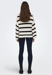 Femme aux longs cheveux bruns portant un pull blanc à rayures horizontales noires, un jean skinny foncé et des bottines noires, vue de dos.