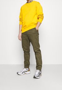 Geel katoenen sweatshirt, olijfgroene chino's en zilveren hardloopschoenen. Het sweatshirt heeft een ronde halslijn; de broek heeft zijzakken.