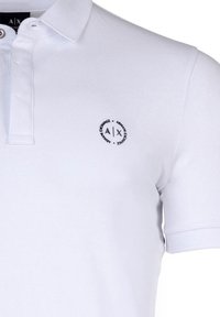 Hvit polo skjorte laget av teksturert stoff, med en to-knapps stolpe og et rundt logo på venstre bryst. Korte ermer med ribbede mansjetter.