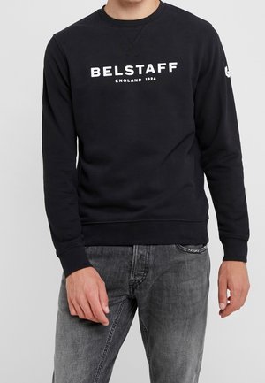 Mężczyzna noszący czarną bluzę Belstaff z białym napisem „BELSTAFF ENGLAND 1924” i wyblakłe szare dżinsy na jednolitym tle.