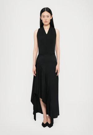 Black, sleeveless dress with a V-neck design. Asymmetrical hemline and soft, flowing fabric. Worn with pointed black shoes.