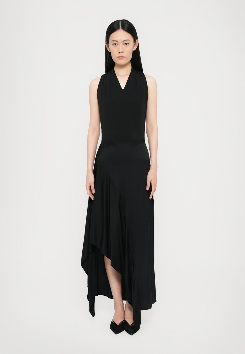 Black, sleeveless dress with a V-neck design. Asymmetrical hemline and soft, flowing fabric. Worn with pointed black shoes.