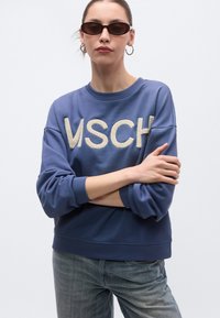 Μπλε φούτερ με το κείμενο "VSCH" σε μπεζ υφασμάτινα γράμματα, με άνετη εφαρμογή και ριμπ μανσέτες, συνδυασμένο με γκρι τζιν και γυαλιά ηλίου.