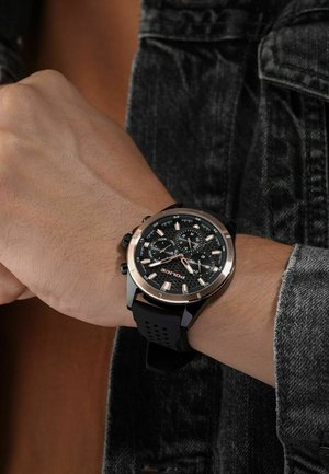 Montre chronographe noire avec un bracelet en silicone noir, des accents en or rose et un cadran noir texturé comportant plusieurs sous-cadrans et des index lumineux.
