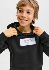 Sudadera negra con bolsillo canguro, que presenta una etiqueta blanca prominente con texto rojo. Tejido suave con puños y dobladillo acanalados.