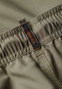 Fascia in tessuto verde con texture arricciata; presenta un'etichetta nera con lettering arancione "Superdry." Dettaglio di cuciture visibili.