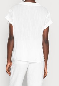 Camicia bianca a maniche corte con una vestibilità rilassata, caratterizzata da un colletto semplice e maniche arrotolate; il tessuto ha un aspetto leggero e leggermente strutturato.