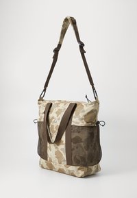 Sac fourre-tout camouflage en beige et marron, avec des panneaux latéraux en mesh, des anses marron et une fermeture à cordon. Design durable et décontracté.