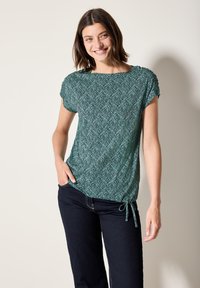 Haut vert à manches courtes avec un motif de feuilles, coupe décontractée, décolleté arrondi, lien ajustable à la taille, associé à un jean foncé.