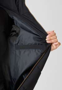 Hand holding open black jacket to show smooth inner lining with a narrow interior pocket and brass-colored zipper on a white background.