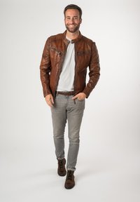 Bruine leren ritsjack met chroomhardware, twee borstzakken, gestructureerde afwerking, gedragen met een lichtgrijze shirt en grijze jeans.