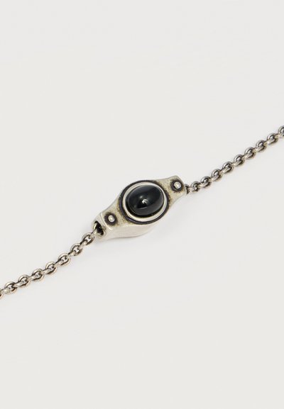 Ασημένιο βραχιόλι με αλυσίδα και οβάλ μαύρη πέτρα τοποθετημένη σε στρογγυλεμένο ορθογώνιο μεταλλικό πλαίσιο με δύο μικρές λεπτομέρειες με καρφιά.