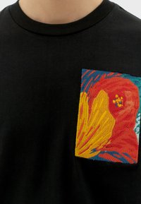 Camiseta de algodón negra con un parche bordado colorido que presenta un diseño abstracto en rojo, amarillo y azul. Mangas cortas y cuello redondo.