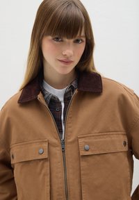 Braune Baumwolljacke mit einem Cordkragen, zwei Fronttaschen und silbernem Reißverschluss. Getragen über einem karierten Hemd mit einem weißen Unterhemd.