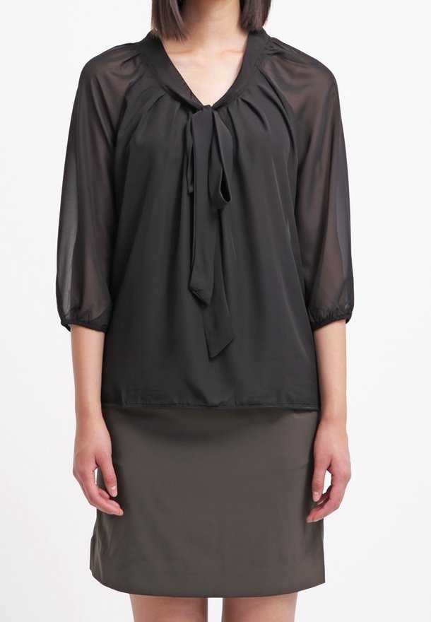 Blouse noire transparente à manches trois-quarts, col en V, avec un détail de noeud à l'encolure, associée à une jupe ajustée vert olive foncé.