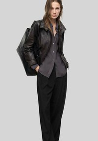 Czarna skórzana kurtka z kołnierzem, szara koszula zapinana na guziki i luźne czarne spodnie. Modelka trzyma czarną torebkę, prezentując warstwowy wygląd.