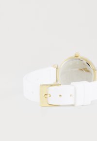 Bracelet de montre en silicone blanc avec une boucle en métal doré à l'extrémité. Le cadran de la montre est rond et dispose d'un couvercle en verre transparent à l'arrière.