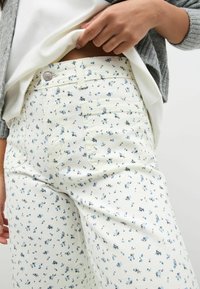 Katoenen broek met bloemenpatroon op een crèmekleurige achtergrond, met kleine blauwe bloemen en zwarte accenten, aansluitend ontwerp en voorzakken.