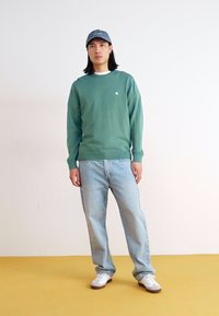 Green knit sweater, light blue jeans, and white sneakers. The sweater features a ribbed neckline and hem; the hat is blue with a logo.