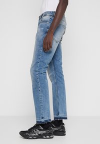 Vaqueros de mezclilla azul claro con corte de pierna recta, con costuras en contraste y un logo en el bolsillo, combinados con zapatos deportivos negros.