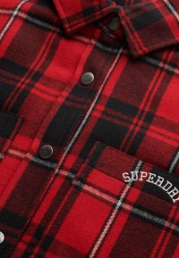 Red and black plaid flannel shirt with silver buttons, featuring a chest pocket and embroidered "SUPERDRY" logo. Soft, textured fabric.
