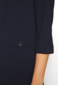 Blouse bleu marine avec manches retroussées, une poche boutonnière argentée, et un tissu texturé pour une finition subtile.