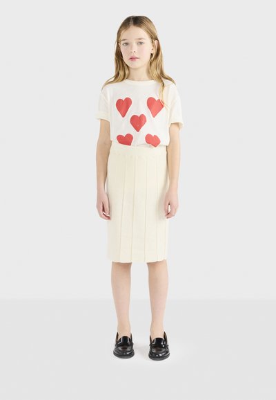Λευκό μπλουζάκι με κόκκινες εκτυπώσεις καρδιάς, συνδυασμένο με κρεμ φούστα με πτυχώσεις. Μαύρα παπούτσια ολοκληρώνουν την εμφάνιση. Απλός και καλαίσθητος σχεδιασμός.
